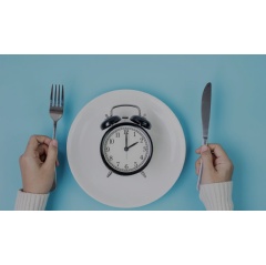 Hands holding knife and fork above alarm clock on white plate on blue background. Intermittent fasting, Ketogenic dieting, weight loss, meal plan and healthy food concept